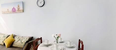 A dining area with a round table set for four, a clock on the wall, and a bed with pillows in the background.