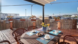 A terrace with wooden tables and chairs, a view of the sea, and a cityscape in the background.