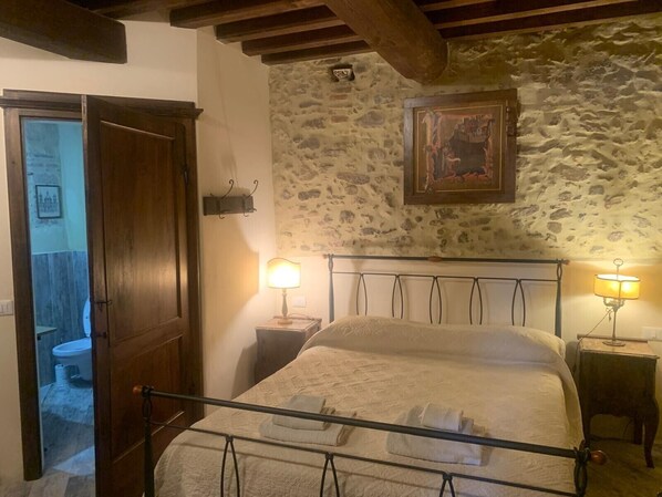 Une chambre avec un mur en pierre, un lit avec un cadre en métal, deux lampes de chevet et une porte donnant sur une salle de bain.