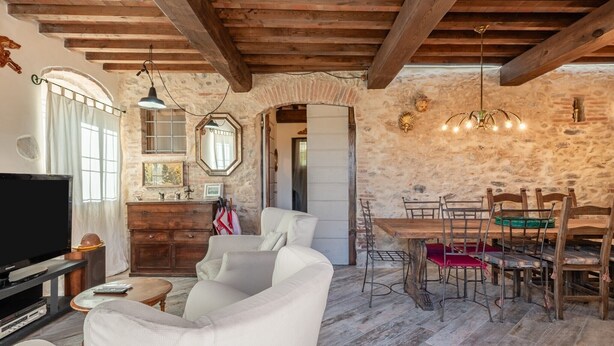 A rustic interior with exposed wooden beams, a stone wall, a dining area with wooden chairs, and a television in the living room. - Holiday Home 'La Capanna Di Arturo Ground Floor' with Mountain View, Wi-Fi and Air Conditioning (Capannori)