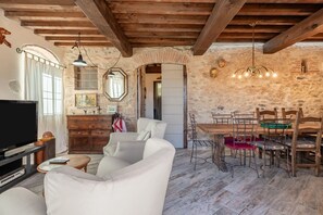 Un interno rustico con travi a vista, una parete in pietra, un'area pranzo con sedie di legno e un televisore nel soggiorno.