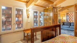 Une chambre avec des poutres en bois, un lit, un banc, une armoire et une vue sur un mur de pierre par la fenêtre.