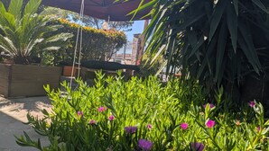 Área de jardim com vasos de plantas e flores, um floreiro de madeira e um guarda-sol vermelho.