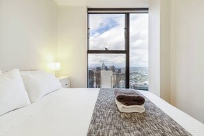 A hotel room with a large window, a bed, a nightstand, and a view of a cityscape.