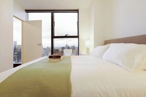 Quarto de hotel com cama, mesa de cabeceira, luminária e uma janela grande com vista para a cidade.