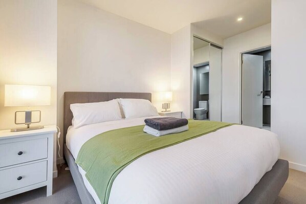 A neatly made bed with pillows, a green throw blanket, and a folded towel. There is a bedside table with a lamp and a small nightstand with a lamp. A mirror is visible on the wall, and a door leads to another room.