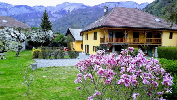 Un bâtiment jaune avec un toit brun, entouré d’un jardin bien entretenu et avec des montagnes en arrière-plan.