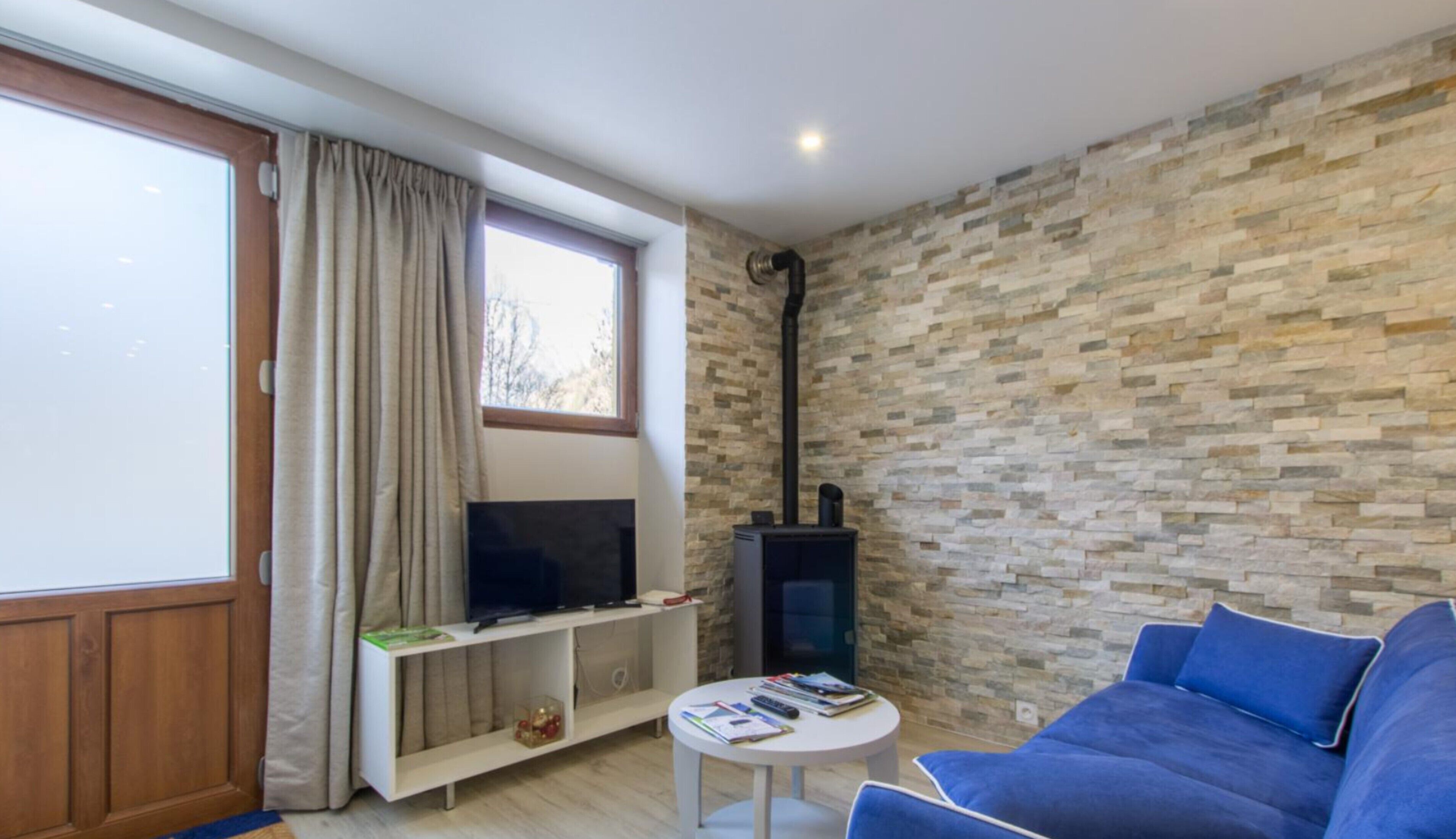 Un salon avec un mur en pierre, un téléviseur posé sur un meuble blanc, un canapé bleu et une petite table avec des livres.