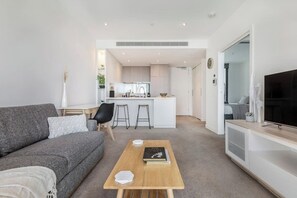 A modern living room with a sofa, a coffee table, and a TV. There is a kitchen area in the background with a dining table and chairs.