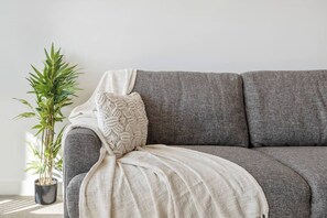 A grey sofa with a white throw blanket and a decorative pillow. A potted plant is visible to the left.