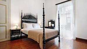 Una cama con dosel de cuatro postes con acabado oscuro, una mesita de noche con lámpara y un amplio ventanal con cortinas translúcidas.