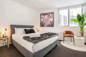 A modern bedroom with a bed, a chair, a plant, and a painting on the wall.