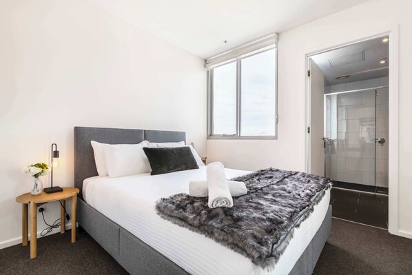 A modern hotel room with a large bed, a bedside table with a lamp, a window, and a shower area.