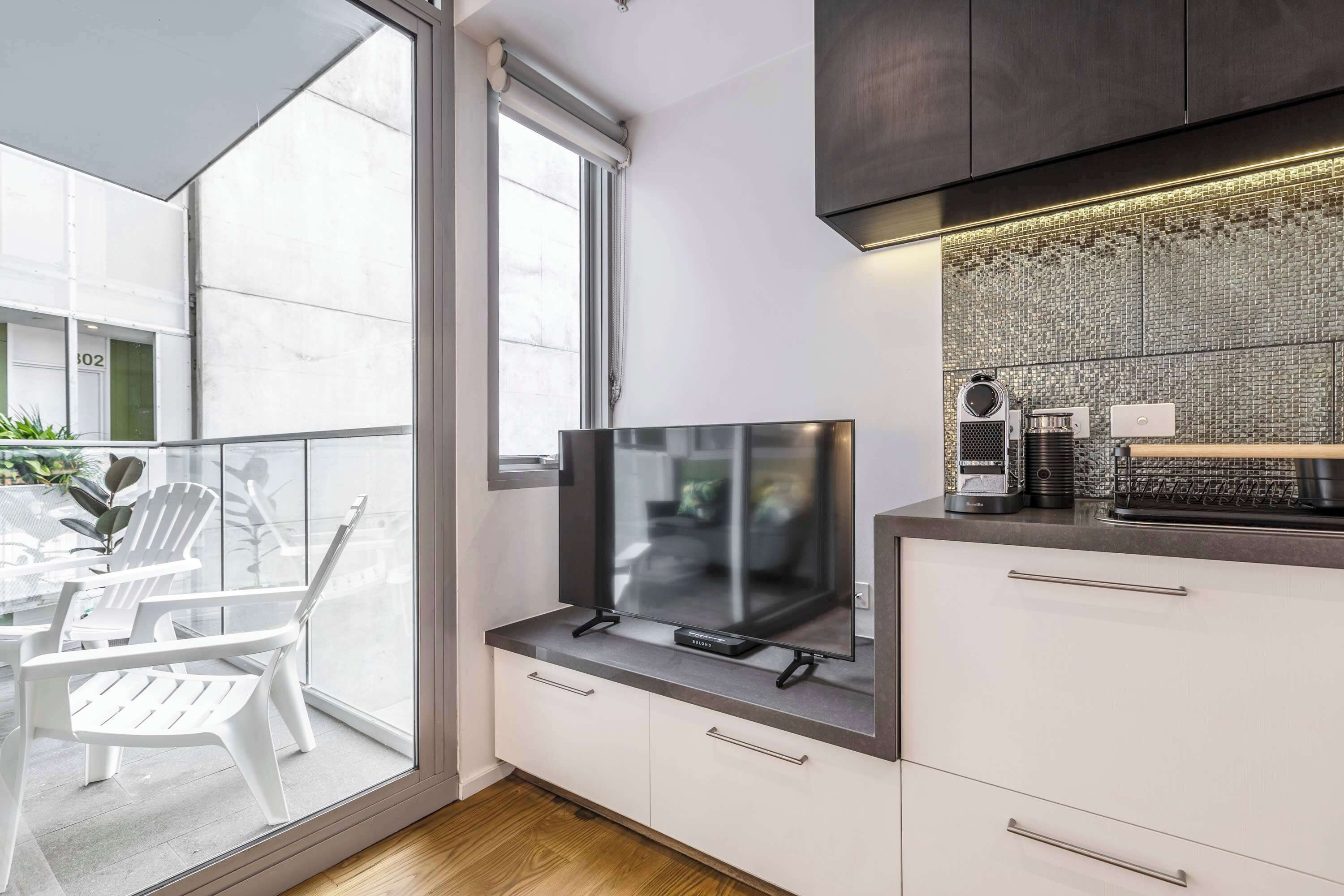 A modern kitchen with a large window, a flat-screen TV, and a small dining area with white chairs.