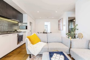A modern kitchen with a built-in oven, a dining area with a gray sofa, and a living room with a white fluffy throw.