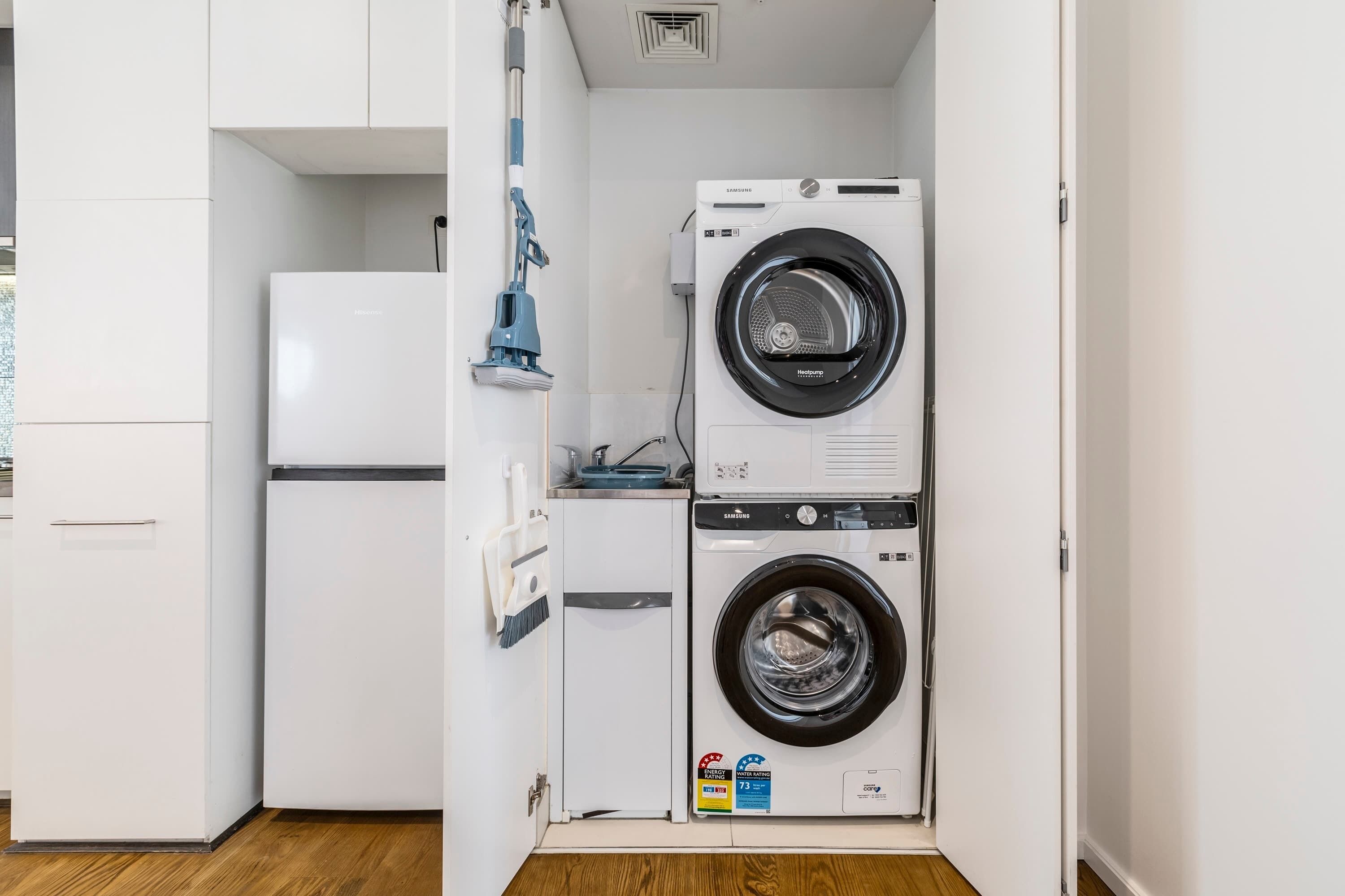 A compact laundry room with a front-loading washer and dryer, a small refrigerator, and a mop.