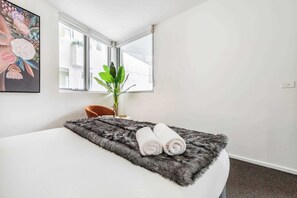 Quarto de hotel com uma cama, uma planta, um quadro e uma janela.