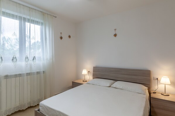 A bedroom with a bed, two bedside tables with lamps, a window with sheer curtains, and wall-mounted light fixtures.