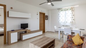 A modern living room with a TV, wooden furniture, a ceiling fan, a dining area, and a colorful sofa. - Apartment 'Ca' Graziosi Cuori' with Mountain View, Shared Garden and Wi-Fi (Bagnone)