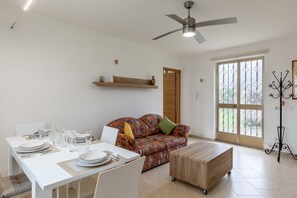 A modern living room with a dining table set, a sofa, a ceiling fan, and a large window.