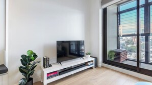 A modern living room with a flat-screen TV, a white entertainment unit, a large window with a city view, and a potted plant.