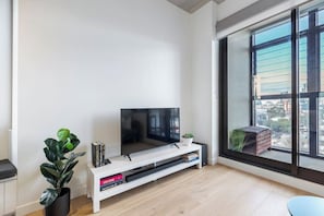 A modern living room with a flat-screen TV, a white entertainment unit, a large window with a city view, and a potted plant.