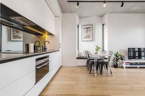 Cozinha moderna com forno embutido, sala de jantar com mesa e cadeiras, e televisão na sala de estar.