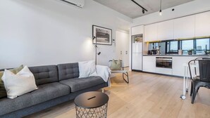 A modern living room with a grey sofa, a small round table, and a kitchen area with white cabinets and a window.