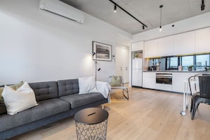 A modern living room with a grey sofa, a small round table, and a kitchen area with white cabinets and a window.