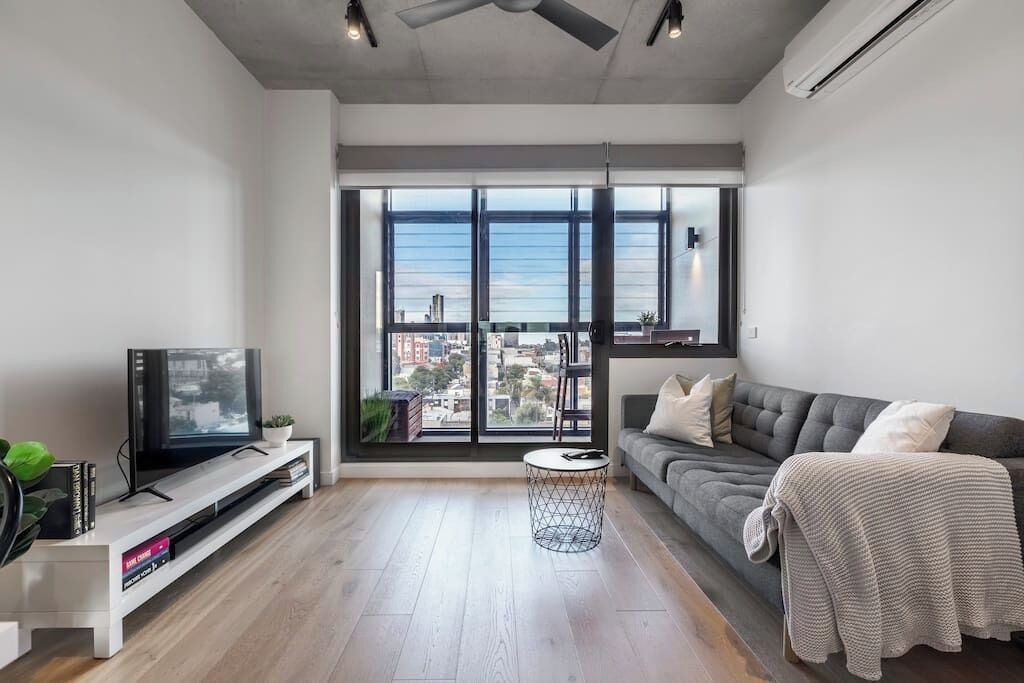 A modern living room with a large window, a grey sofa, a TV on a white stand, and a small round table.