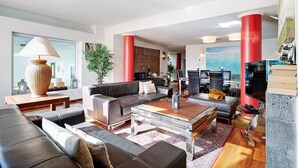 Een moderne woonkamer met een bank, salontafel en televisie. Er zijn rode zuilen en een groot raam.