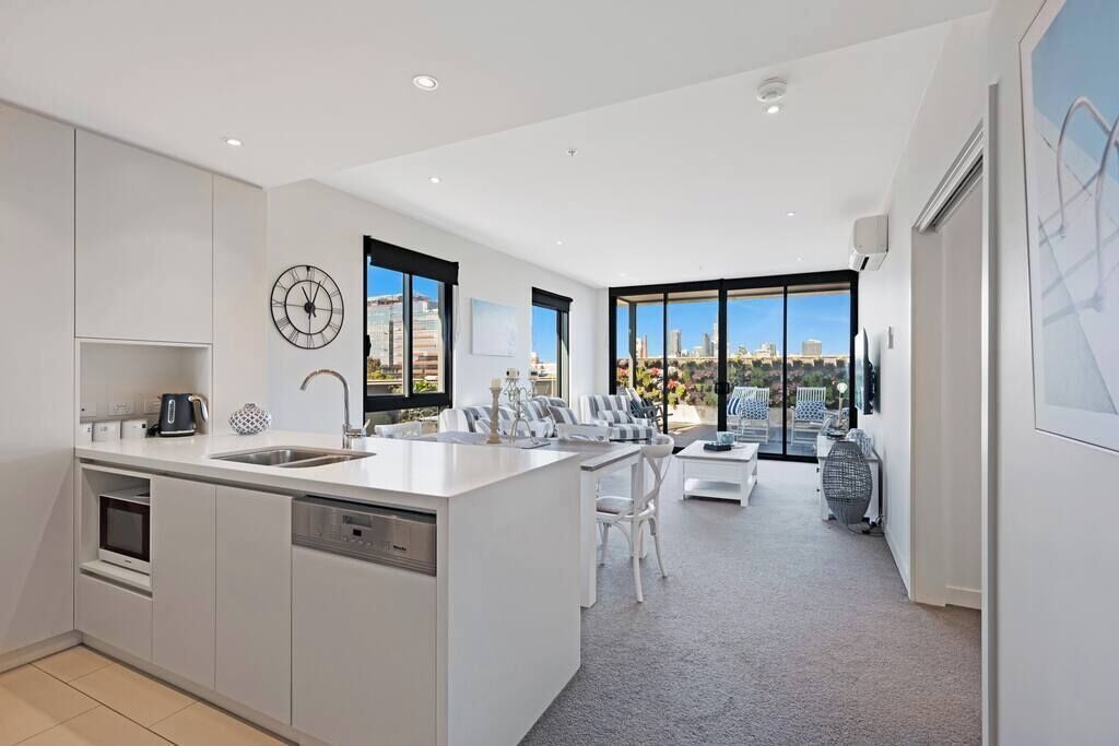 A modern kitchen with a central island, a dining area with white chairs, and large windows offering a city view.