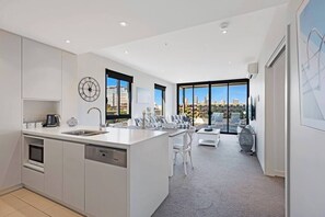 A modern kitchen with a central island, a dining area with white chairs, and large windows offering a city view.