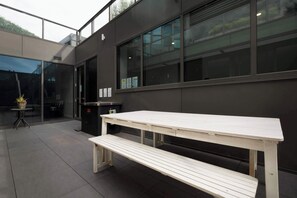 A modern outdoor seating area with a long white table and bench, a glass door entrance, and a view of the building's exterior.