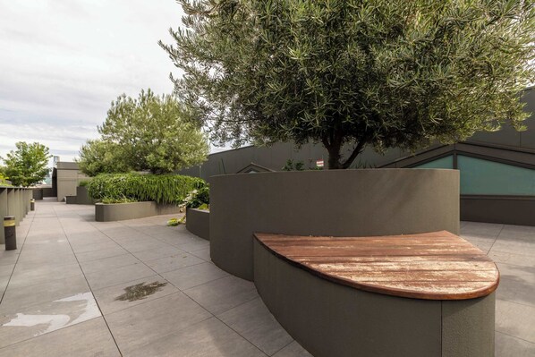 A courtyard with a curved wooden bench, planters with greenery, and a large tree.