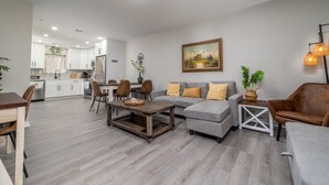 A modern living room with a sofa, coffee table, and chairs. There is a kitchen in the background with white cabinets and a sink.