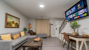 A living room with a grey sofa, wooden coffee table, and a TV mounted on the wall displaying various channels.