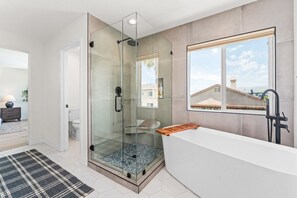 A modern bathroom with a glass-enclosed shower, a freestanding bathtub, and a large window offering a view of buildings.