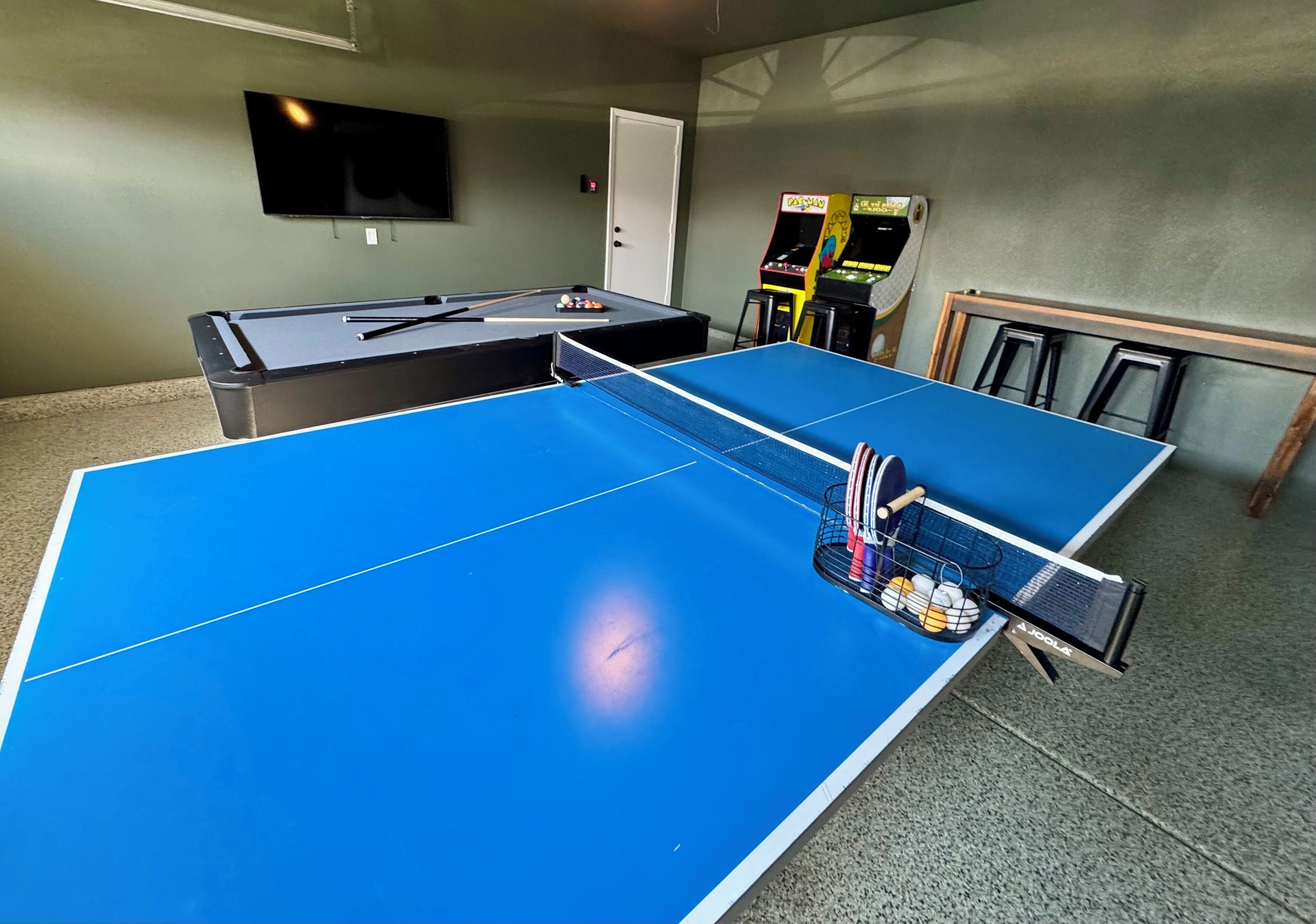 Una mesa de ping pong con una red en el centro, una mesa de billar con bolas, y un televisor montado en la pared.
