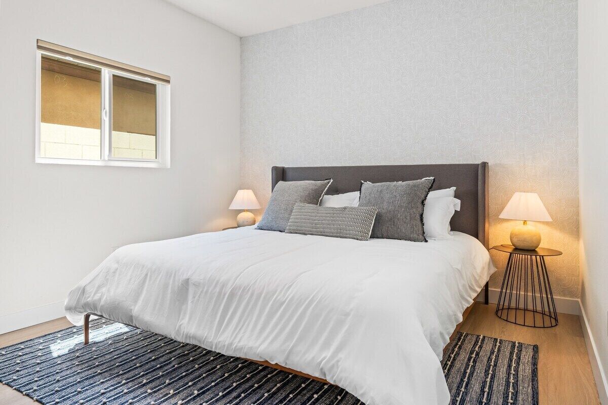 A neatly made bed with white bedding, two bedside lamps, and a patterned rug on a wooden floor.