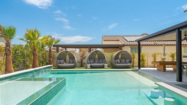 A swimming pool with three hanging chairs, palm trees, and a building with a tiled roof. - Green Lion - New Spectacular Modern Pool home (Coachella)
