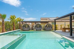 A swimming pool with three hanging chairs, palm trees, and a building with a tiled roof.