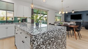 A modern kitchen with a large island, white cabinets, and a green backsplash. There is a dining area with a table and chairs, and a TV mounted on the wall. - Green Lion - New Spectacular Modern Pool home (Coachella)