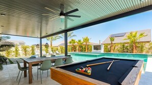A pool table with a pool cue and balls, a ceiling fan, and a view of a swimming pool and palm trees. - Green Lion - New Spectacular Modern Pool home (Coachella)