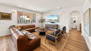 A modern living room with a brown leather sofa, a round coffee table, and a wooden dining table with chairs. There is a TV mounted on the wall and a painting above it. - Los Arcos- 7 bedrooom, sleeps 19 Pool Home (Coachella)