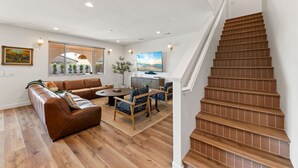 A modern living room with a brown leather sofa, a round coffee table, and blue patterned chairs. A wooden staircase leads to a landing with a painting and a view of the outdoors. - Los Arcos- 7 bedrooom, sleeps 19 Pool Home (Coachella)