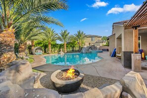 A backyard with a swimming pool, a fire pit, palm trees, and a patio area.