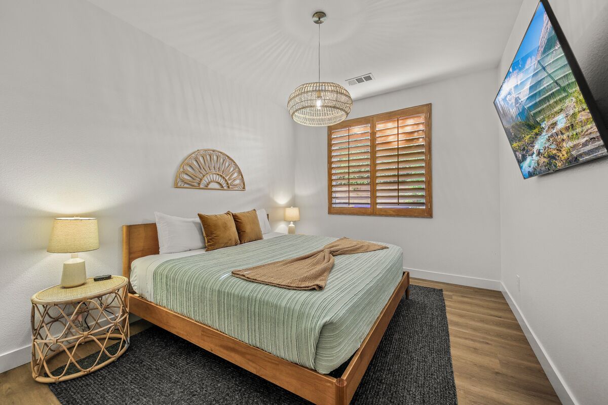 A bedroom with a wooden bed, a round wicker side table, a wall-mounted lamp, a decorative wall piece, a window with blinds, and a framed picture on the wall.