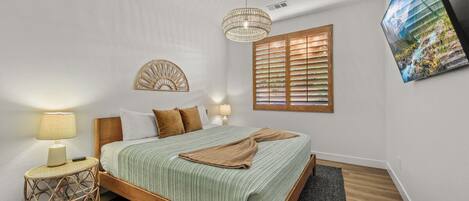 A bedroom with a wooden bed, a round wicker side table, a wall-mounted lamp, a decorative wall piece, a window with blinds, and a framed picture on the wall.