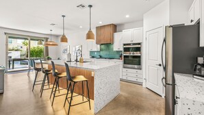 A modern kitchen with a central island, bar stools, a large window, and a view of greenery outside. - Escape to this newly remodeled 7-bedroom desert retreat designed for relaxation and fun. Enjoy a large resort-style pool and spa, a fully stocked kitchen, spacious game room, and outdoor games perfect for group play under endless blue skies. Whether (Coachella)